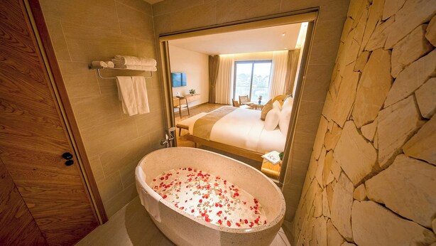 Een badkamer met een stenen muur, een badkuip met rozenblaadjes, een houten deur en een spiegel die een slaapkamer weerspiegelt.