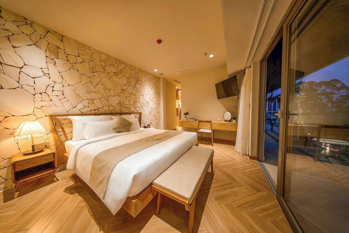 Un dormitorio moderno con pared de piedra, una cama grande, una mesita de noche, un banco, un escritorio y vistas a través de un ventanal.
