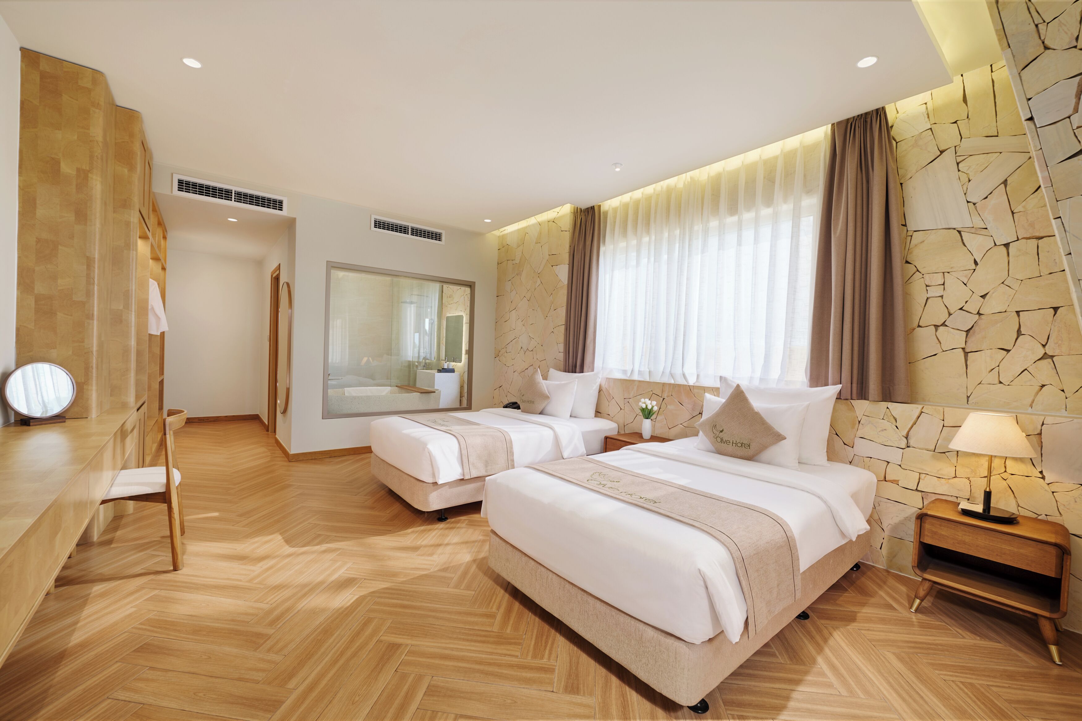 A hotel room with two beds, a sitting area with a chair and table, a large mirror, and a stone-patterned wall.