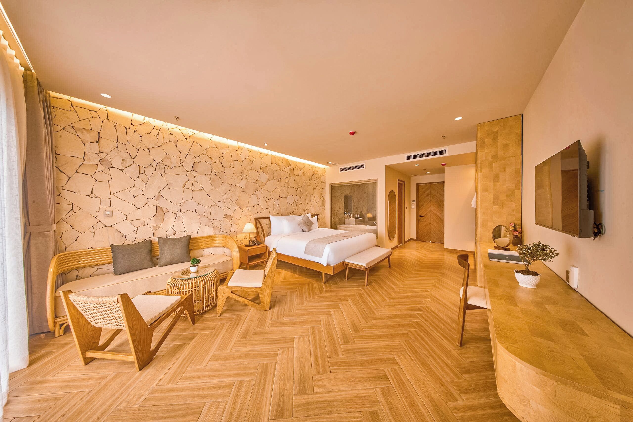Una camera d'albergo moderna con parete in pietra, un letto, una poltrona, un tavolino in legno, una televisione e una pianta.