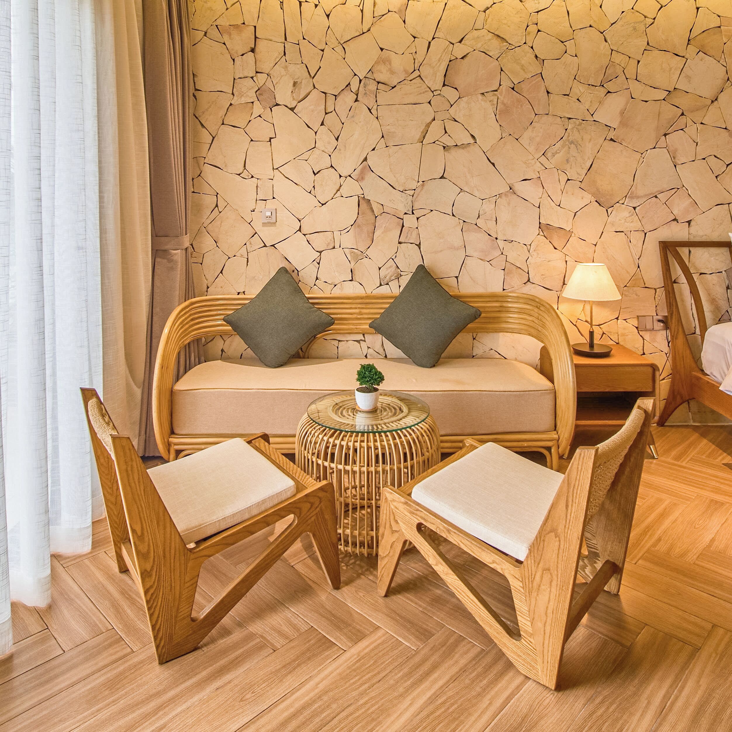 A room with a stone wall, a sofa with cushions, two wooden chairs, a small table with a plant, and a lamp on a wooden nightstand.