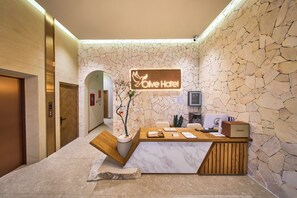 Un comptoir d’accueil avec un mur en pierre en arrière-plan, une plante en pot et une enseigne indiquant « Hôtel Olive ».