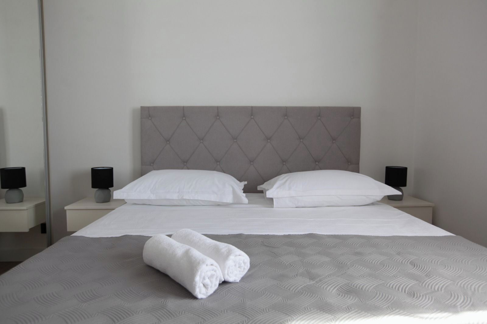 A neatly made bed with white linens and two rolled towels, a tufted headboard, and bedside lamps on nightstands.