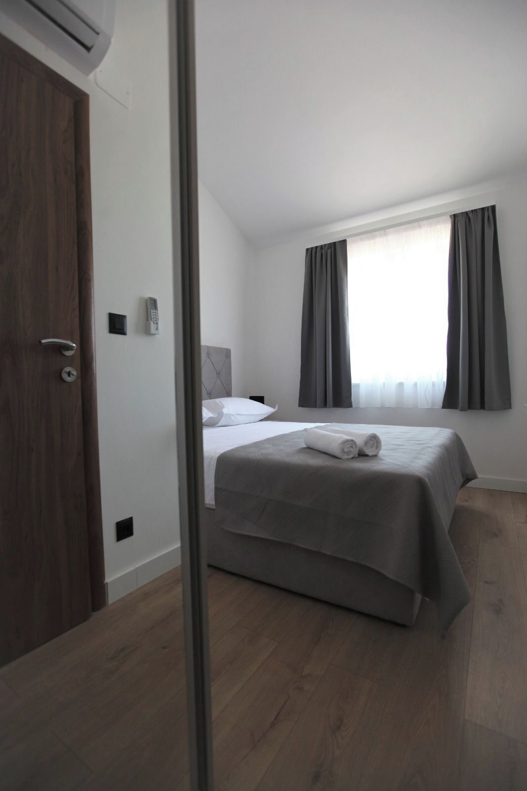 Habitación de hotel con cama, mesita de noche, ventana con cortinas, puerta y equipo de aire acondicionado.