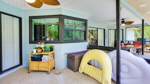 Een kamer met een groot raam, plafondventilatoren, een rieten karretje met planten en een opblaasbaar glijbaantje.