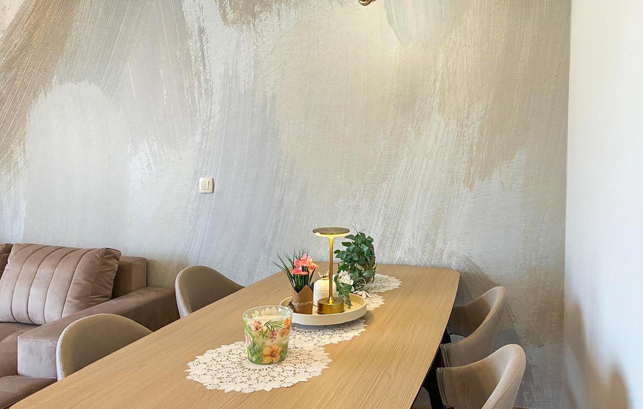 A dining area with a wooden table, chairs, a small round table with a lamp, and a vase with flowers.