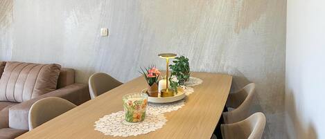 A dining area with a wooden table, chairs, a small round table with a lamp, and a vase with flowers.