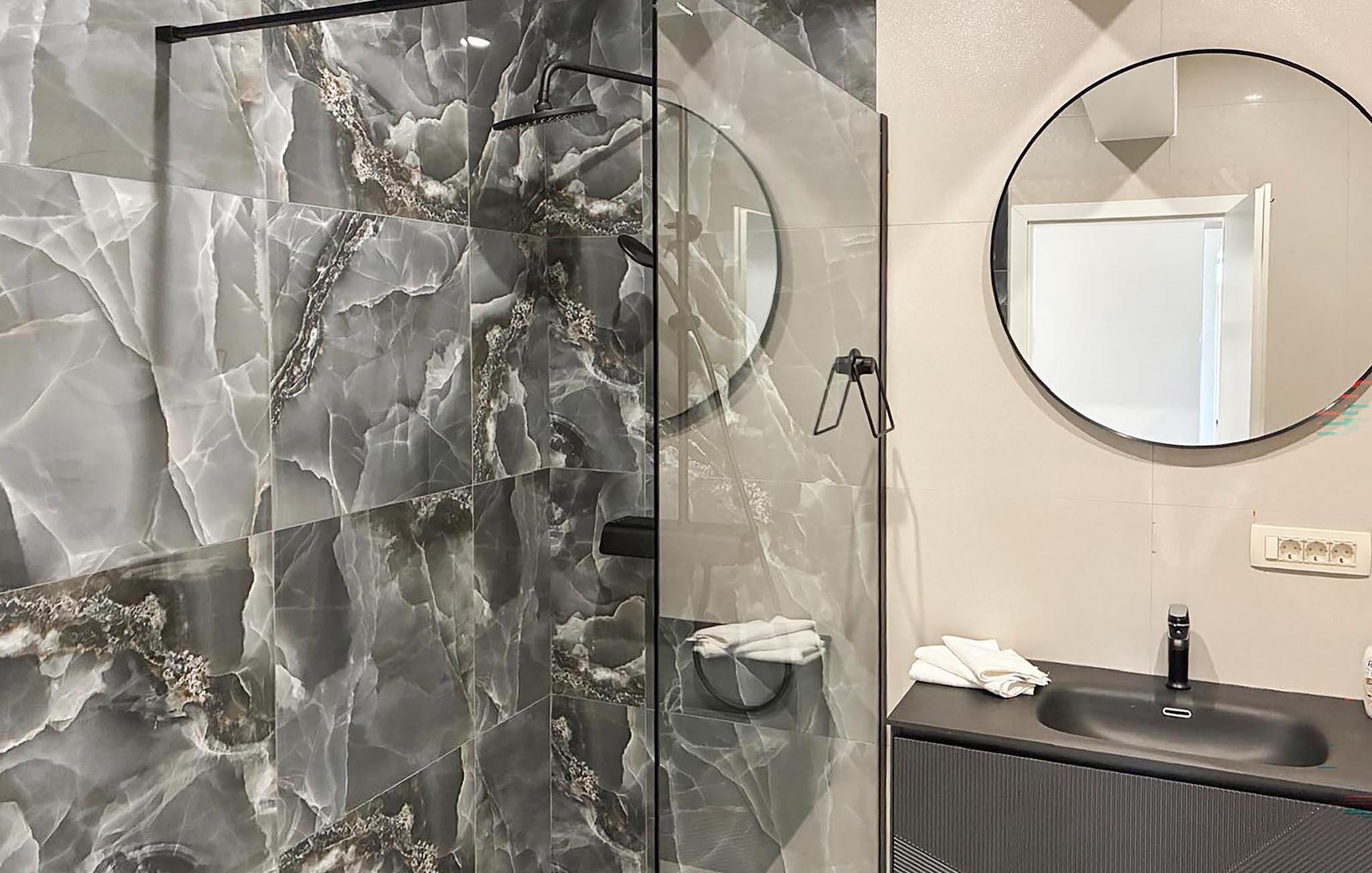 A bathroom with a glass shower enclosure, a round mirror, a sink, and a towel rack.