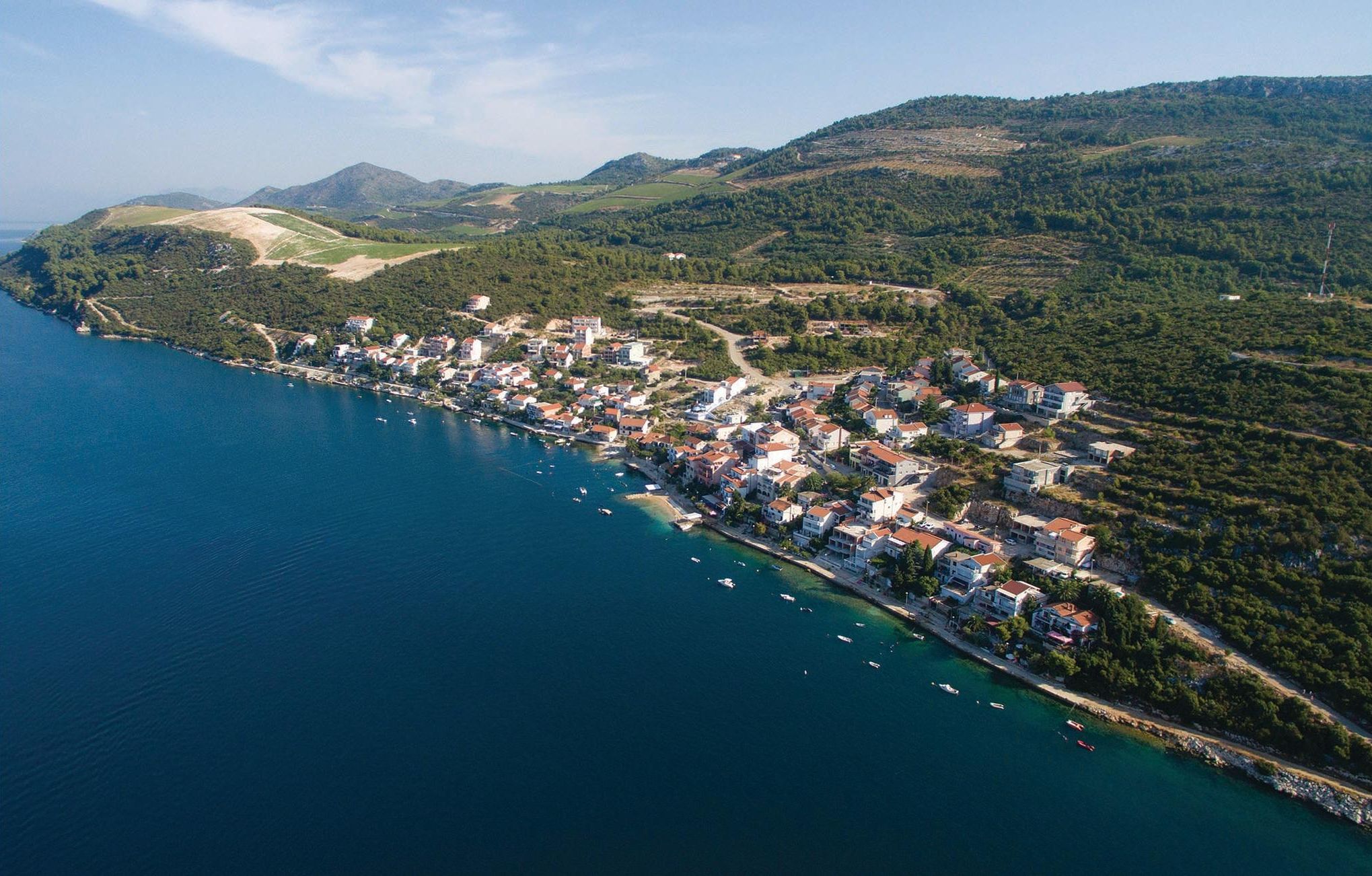 An aerial view of a coastal town with buildings along the shoreline, a clear blue sea, and hilly terrain in the background.