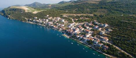 An aerial view of a coastal town with buildings along the shoreline, a clear blue sea, and hilly terrain in the background.