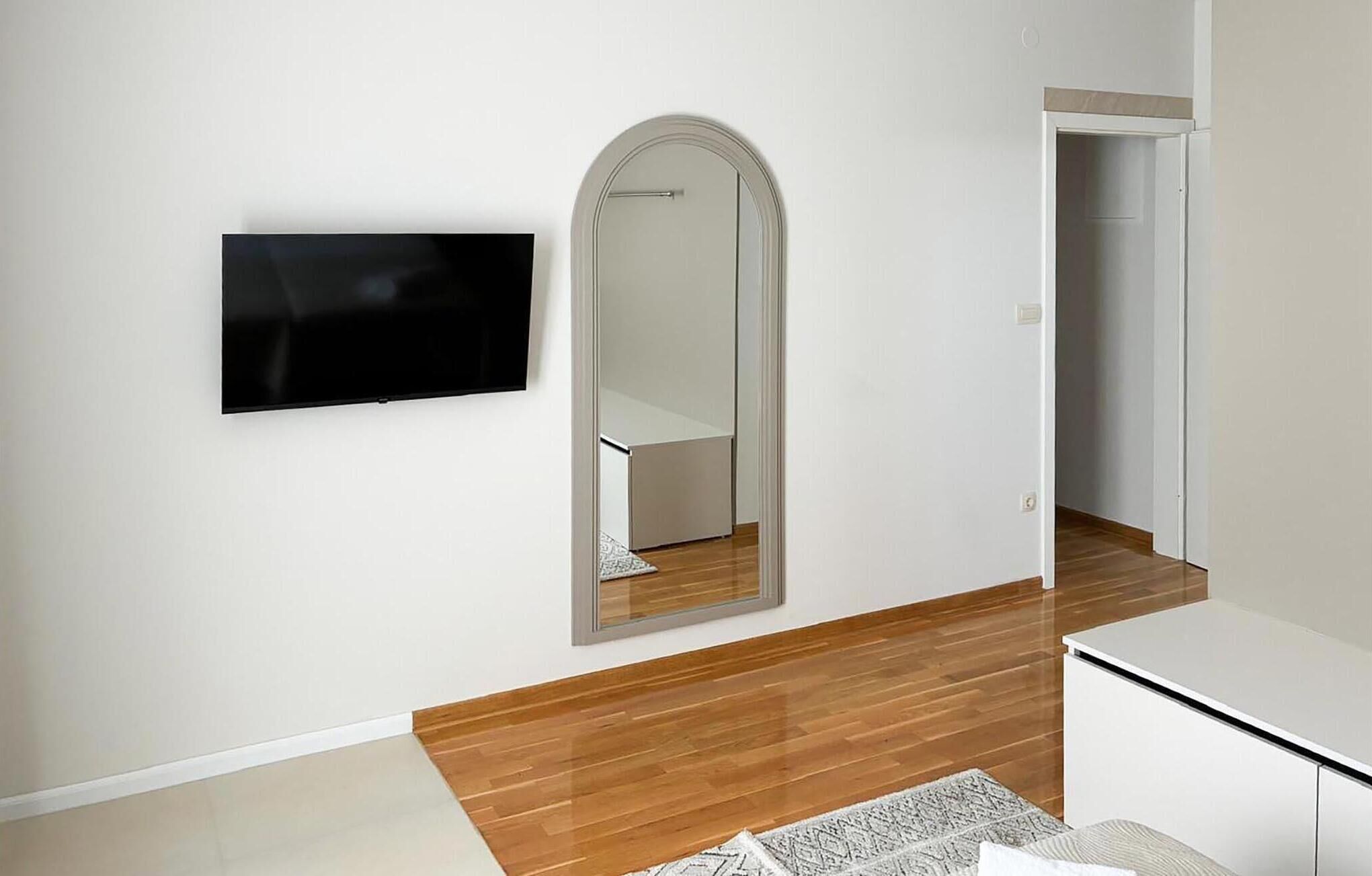Une pièce dotée d’un téléviseur à écran plat fixé au mur, d’un grand miroir, d’un plancher en bois et d’un bureau blanc.