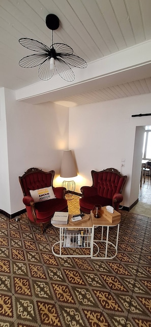 Ett rum med ett mönstrat golv, två röda fåtöljer, ett bord med en lampa, en takfläkt och en dörr som leder till ett annat rum.