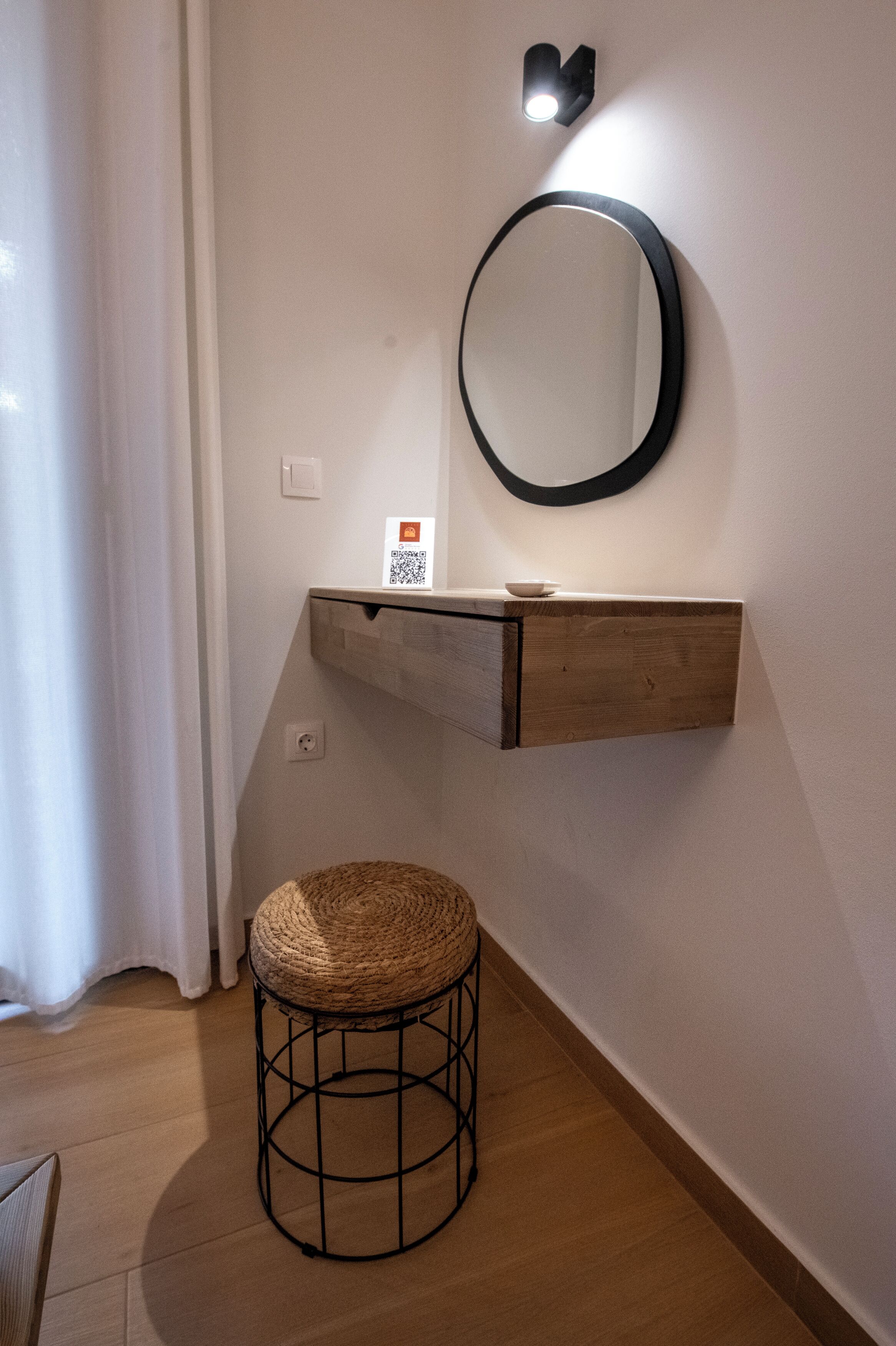 Ein Badezimmer mit einem Holzinlay, einem runden Spiegel, einem Schild mit einem QR-Code und einem gewobenen Hocker.