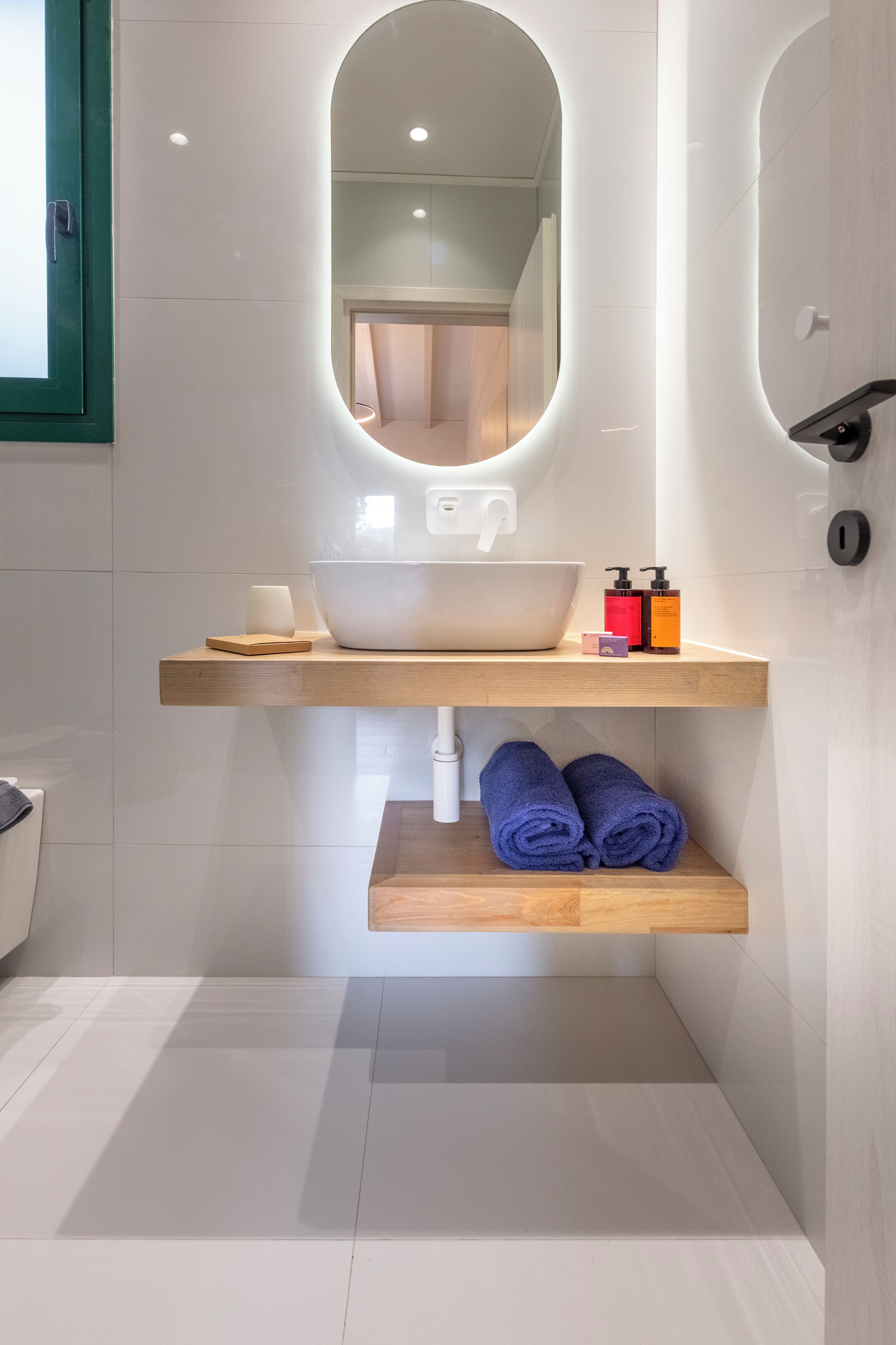Une salle de bain avec un lavabo blanc, un miroir et une étagère en bois où sont posées des serviettes.