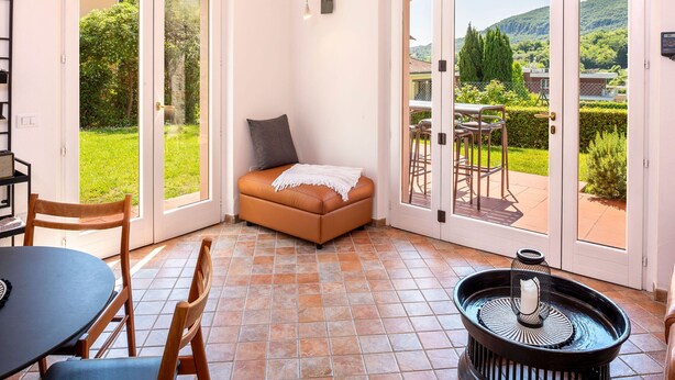Ein Zimmer mit einer Couch, einem Tisch, Stühlen, einem kleinen Tisch mit einer Kerze und Blick durch Glastüren auf einen Außenbereich mit Rasen und Stühlen.