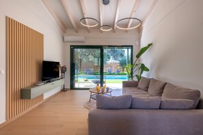 Ein modernes Wohnzimmer mit einer Couch, einem Fernseher auf einer Holzvertäfelung, einem Esstisch mit Tassen und Blick auf Pool und Garten durch Glastüren.