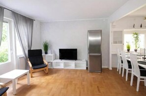Ein modernes Wohnzimmer mit Fernseher, einem Essbereich mit weißen Stühlen, einem Kühlschrank und einem Fenster mit Vorhängen.