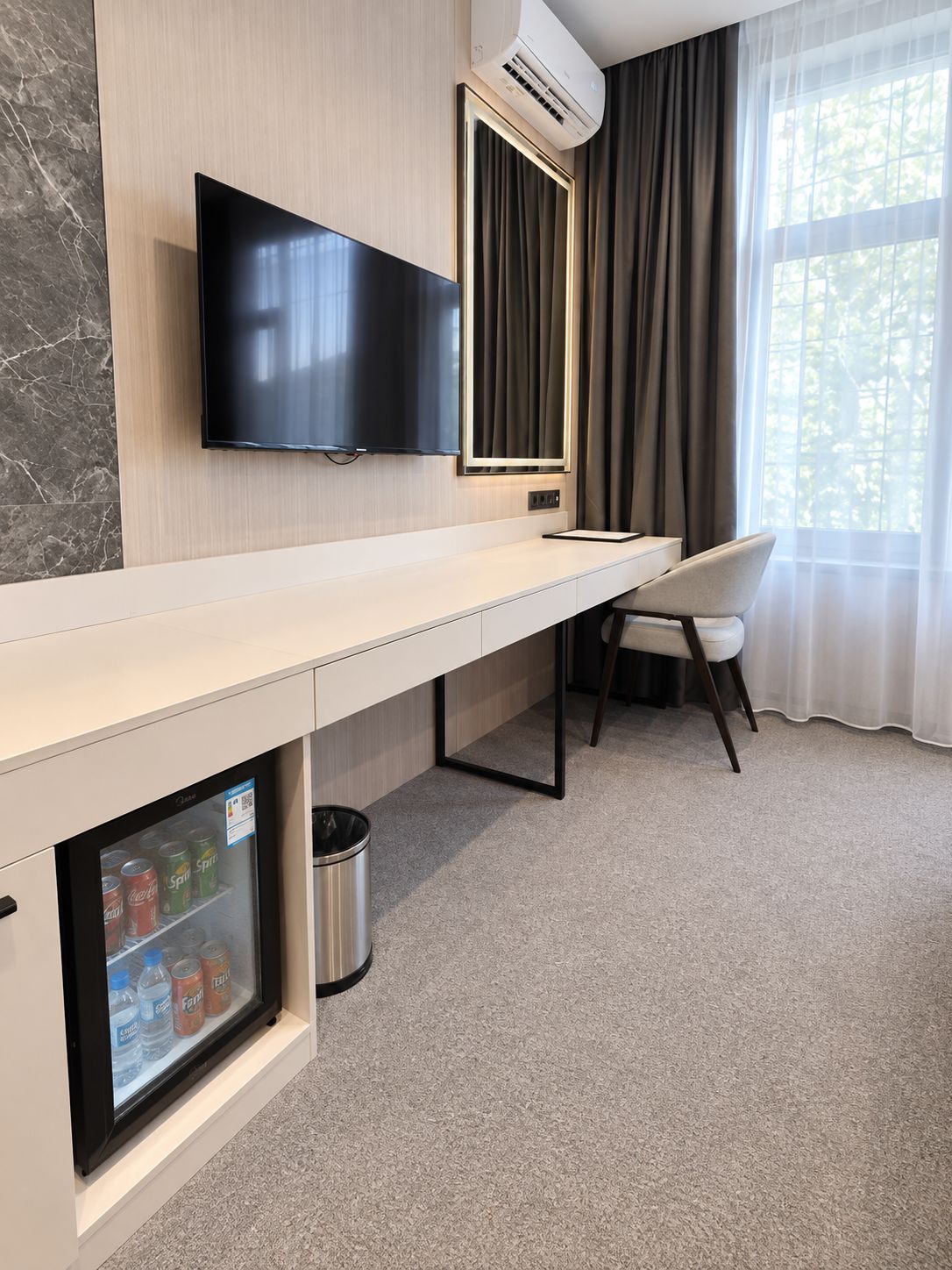 A hotel room with a flat-screen TV mounted on the wall, a minibar with beverages, a trash can, a chair, and a window with curtains.