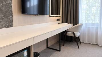 A hotel room with a flat-screen TV mounted on the wall, a minibar with beverages, a trash can, a chair, and a window with curtains.