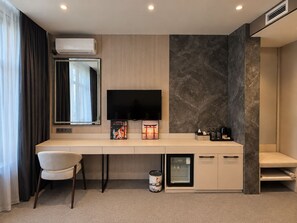 A modern hotel room with a flat-screen TV, a small refrigerator, a coffee machine, a trash can, and a desk with a chair. - Tokyo Hotel (Bishkek)