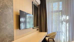 A modern room with a flat-screen TV mounted on the wall, a wooden desk, a chair, and a large window with sheer curtains. - Tokyo Hotel (Bishkek)