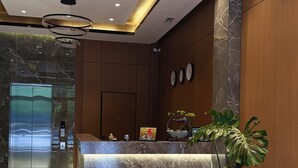 A hotel reception area with a marble counter, a large potted plant, a modern chandelier, and a digital elevator display. - Tokyo Hotel (Bishkek)