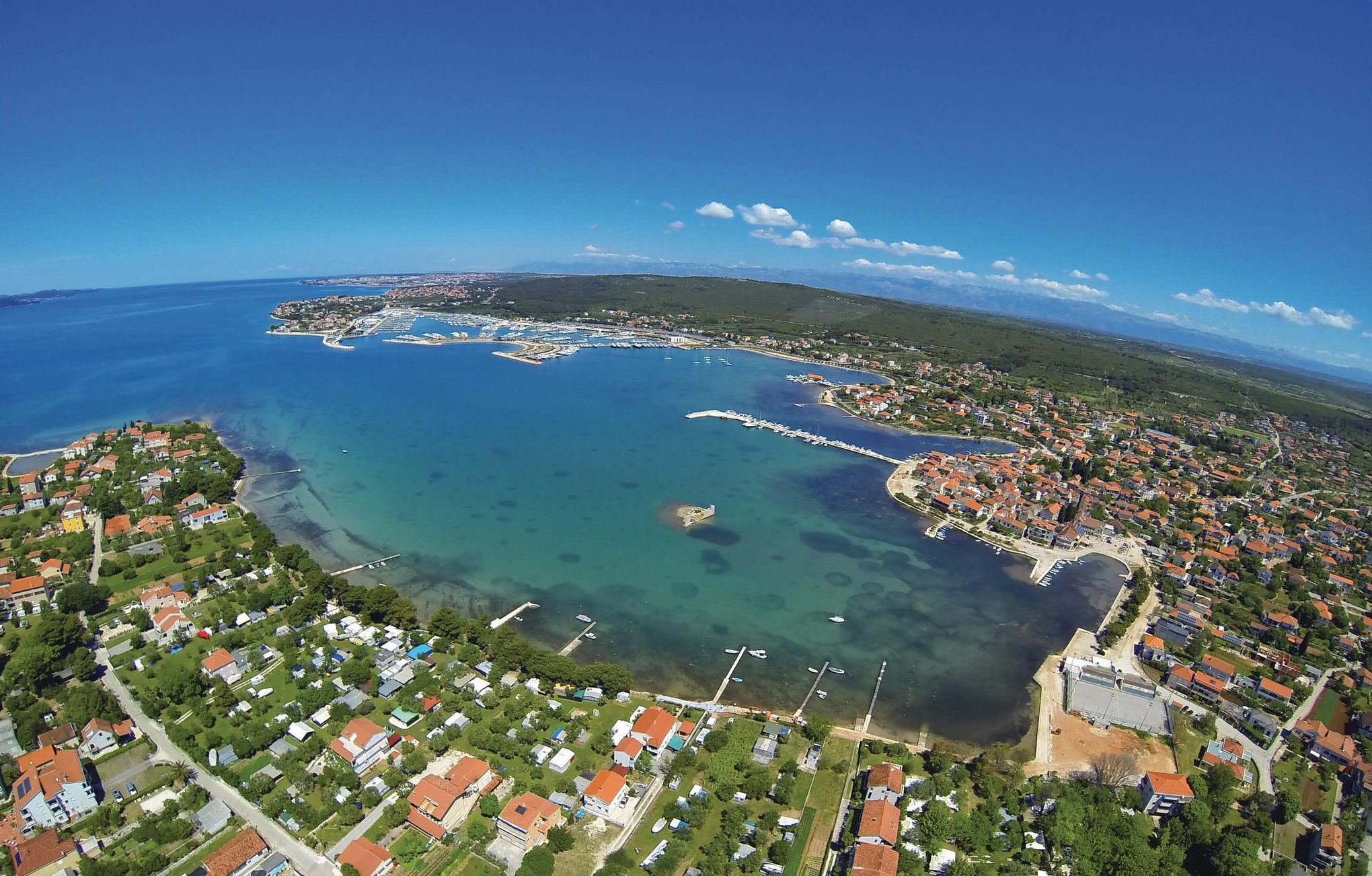 Vista aérea de uma vila costeira com águas cristalinas, um porto de iates e áreas residenciais.