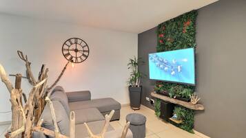 A modern living room with a grey sofa, a wall-mounted TV, a clock, and decorative plants.