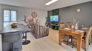Un espace repas moderne avec une table en bois, des chaises en osier, un téléviseur et une composition florale décorative.