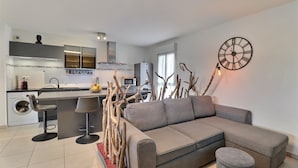 Un salon moderne avec un canapé d’angle gris, une horloge ronde fixée au mur et un coin cuisine à l’arrière-plan.