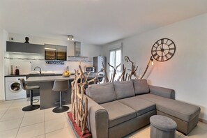 A modern living room with a grey sectional sofa, a round wall clock, and a kitchen area with a washing machine. - Evasion Tropicale (Ajaccio)