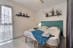 A neatly made bed with a blue blanket, white pillows, and folded towels. There are two wall-mounted shelves with decorative items. - Evasion Tropicale (Ajaccio)