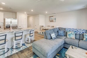 Eine moderne Küche mit Einbaukühlschrank, ein Essbereich mit Tisch und Stühlen sowie ein Wohnzimmer mit grauem Ecksofa und gemusterten Kissen.