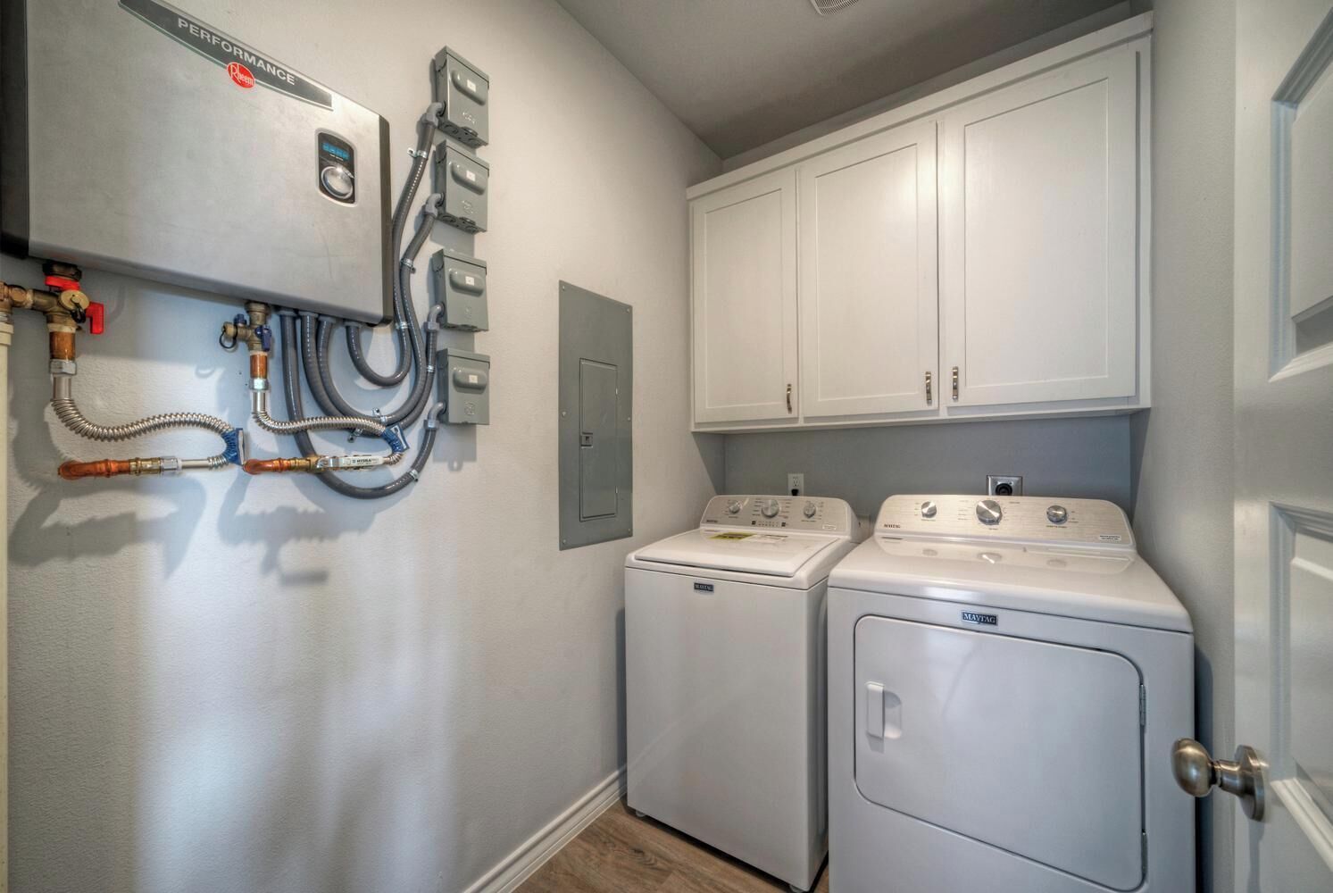 A laundry room with two front-loading washing machines, a dryer, and a wall-mounted control panel with hoses and valves.