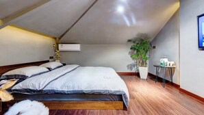 Ein Schlafzimmer mit schrägverdachter Decke, einem Bett mit Holzrahmen, einer Topfpflanze und einem kleinen Tischchen mit Wasserkocher.