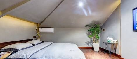A bedroom with a sloped ceiling, a bed with a wooden frame, a potted plant, and a small table with a kettle.