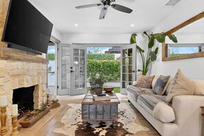 A living room with a stone fireplace, a flat-screen TV, a ceiling fan, a sofa, and a large window. - Designer Home + Backyard Oasis + Firepit | Mins to Universal | Laurel Terrace (Los Angeles)