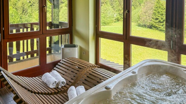 Ein Wellnessraum mit einer Holzbank, Handtüchern, einer Whirlwanne und großen Fenstern mit Blick auf den Wald.