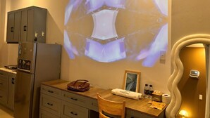 Habitación de hotel con kitchenette, un escritorio de madera, una silla, un proyector montado en la pared y un cuadro enmarcado.