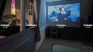 Una habitación con una pantalla grande que muestra un paisaje urbano y un artista, una ventana con vistas a un edificio alto, y el texto 'Darlington HOUSE' en la pantalla.