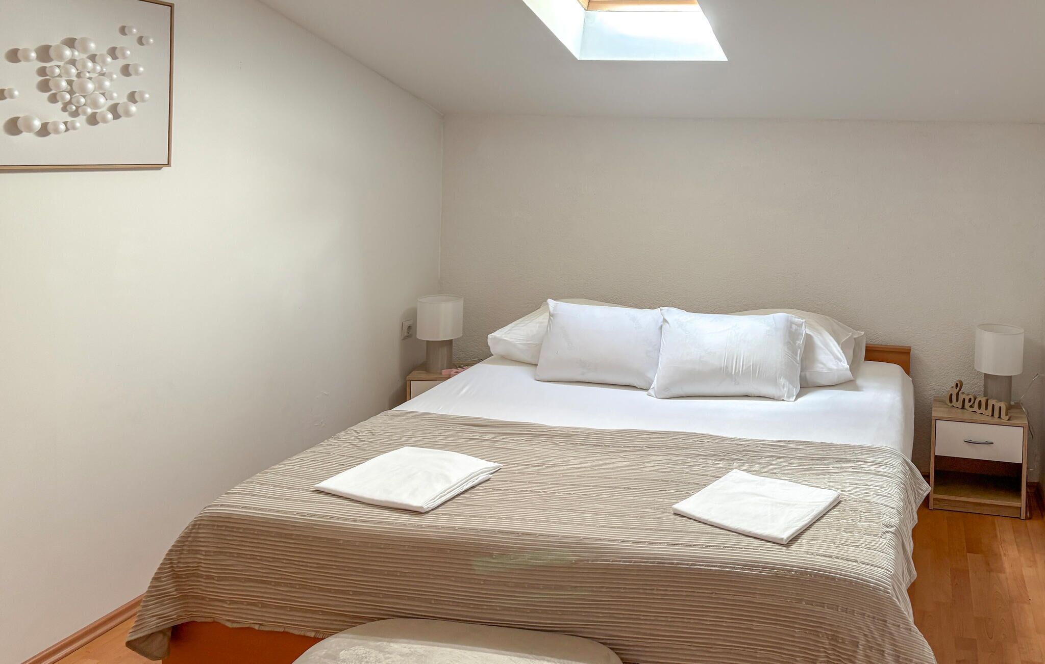 A neatly made bed with white linens, a striped blanket, and two pillows. There are two bedside tables with lamps and a small wooden nightstand with a lamp and a decorative item. A framed artwork is on the wall above the bed.