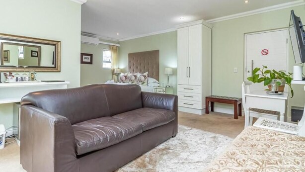 A bedroom with a brown leather sofa, a bed with a patterned bedspread, a white wardrobe, a small bedside table, and a mirror on the wall. - Hermanus Cliff Path Lodge (Hermanus)