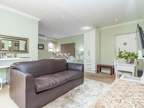 A bedroom with a brown leather sofa, a bed with a patterned bedspread, a white wardrobe, a small bedside table, and a mirror on the wall. - Hermanus Cliff Path Lodge (Hermanus)