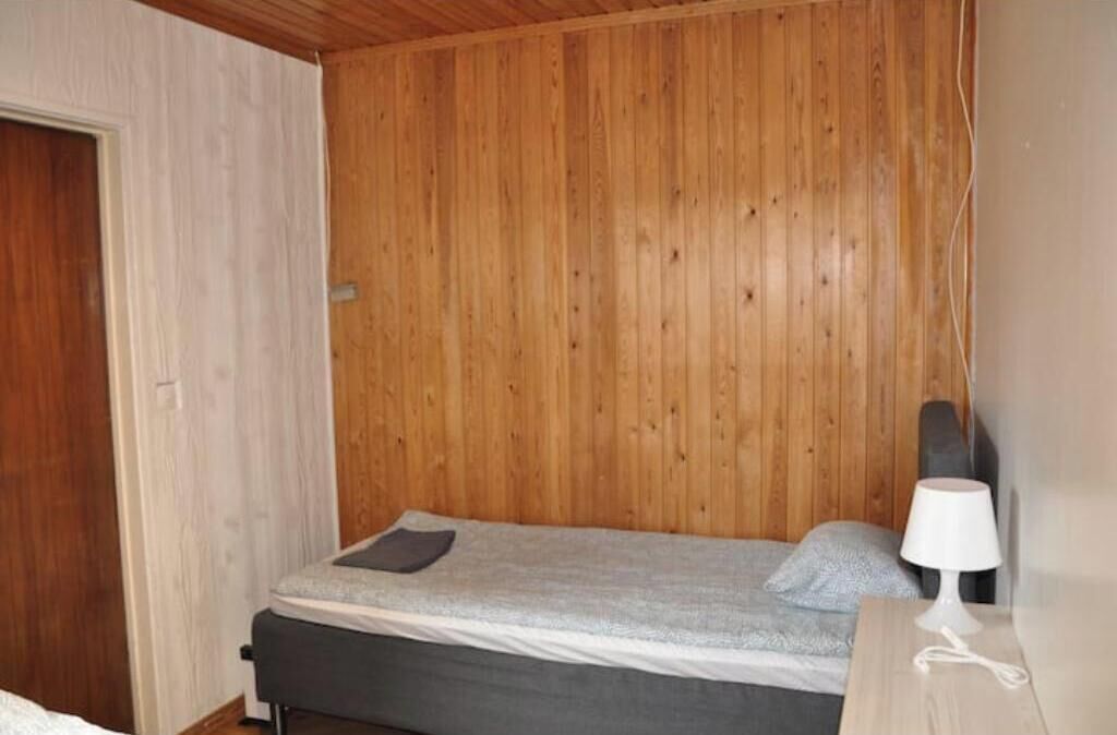 Une petite chambre avec un lit simple, des lambris en bois sur les murs, une table de chevet avec une lampe et une porte sur le côté gauche.