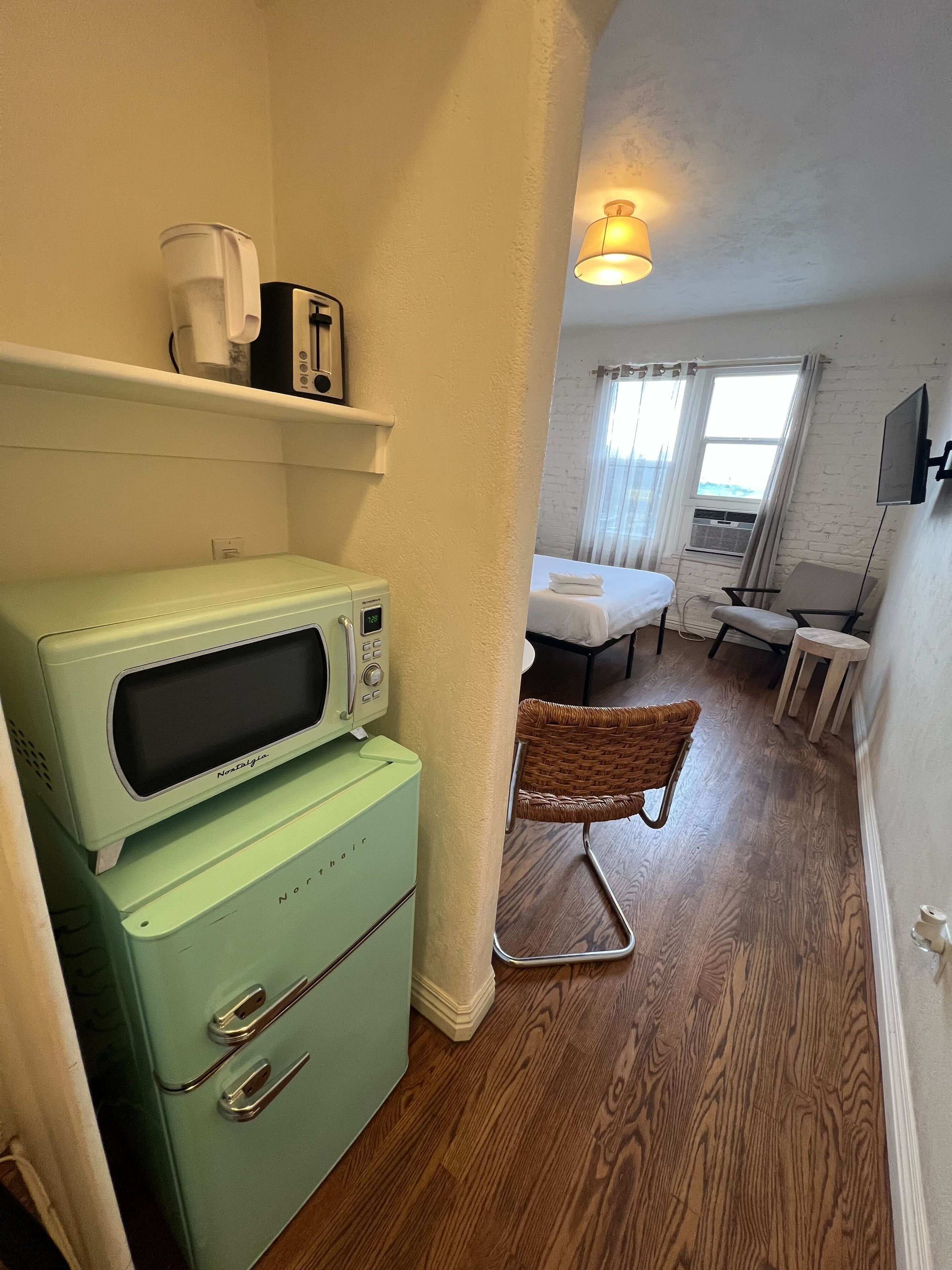 A compact hotel room with a kitchenette, a bed, a television, and a window with curtains.