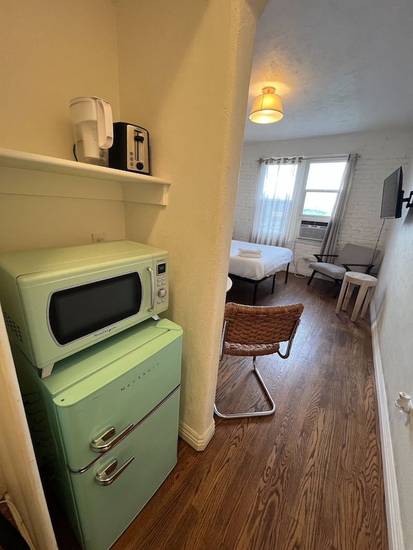 A compact hotel room with a kitchenette, a bed, a television, and a window with curtains.