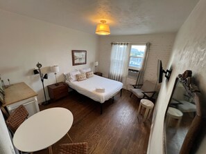 A hotel room with a bed, bedside tables, a desk, a chair, a dining table, and a window with curtains.