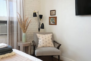 A hotel room with a bed, a chair, a small table, a vase with dried plants, a floor lamp, and two framed pictures on the wall.