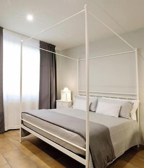 A four-poster bed with a white frame, a bedside table with a lamp, and a view of the outside through a window with curtains. - The Seven Ways - Maple Apartment with Jacuzzi and Large Porch (Piemonte)