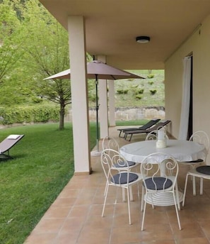 A covered patio with a round table and chairs, a sun lounger, and a view of a grassy area with trees. - The Seven Ways - Maple Apartment with Jacuzzi and Large Porch (Piemonte)